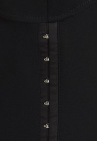 Vêtement en tissu noir avec une surface texturée, comportant six fermetures en métal argenté le long du devant pour la fermeture.