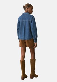 Chaqueta de mezclilla azul con mangas largas, detalle en el yugo trasero, combinada con pantalones cortos de ante marrón y botas de ante marrón hasta la rodilla.