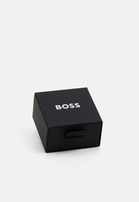 Musta neliön muotoinen laatikko, jossa on mattapintainen pinta. Laatikossa on vetoketju ja sen päällä lukee vahvoilla valkoisilla kirjaimilla "BOSS".