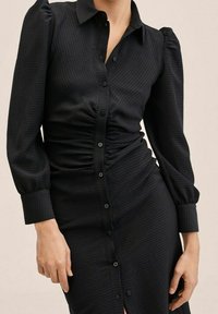 Schwarzes Knopfkleid aus strukturiertem Stoff, mit langen Puffärmeln und gerafften Vorderdetails, in knielanger Schnittführung und mit dezentem Muster.
