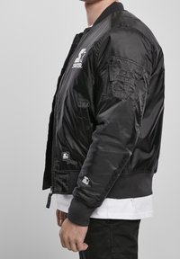 Giacca bomber nera con finitura in satin, colletto e polsini a coste, tasche laterali e patch del logo sulla manica. Presenta una chiusura frontale con zip.