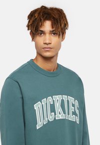 Grön sweatshirt med rundad halsringning, med "DICKIES" i vitt och ljusgrönt broderat tryck över bröstet. Slät textur.