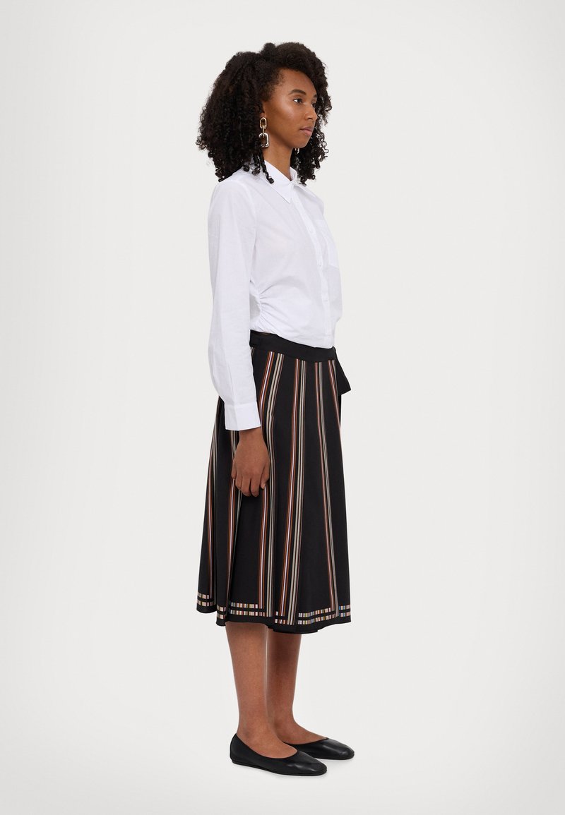 Camisa blanca de botones metida dentro de una falda negra con rayas verticales coloridas. Pendientes cuadrados y bailarinas negras completan el atuendo.