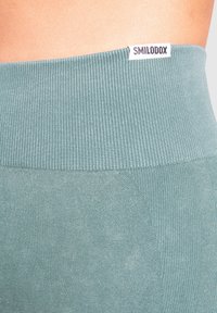 Leggings verde a vita alta con un elastico a coste in vita, dotati di una etichetta con logo. Il tessuto ha un aspetto liscio e texturizzato.