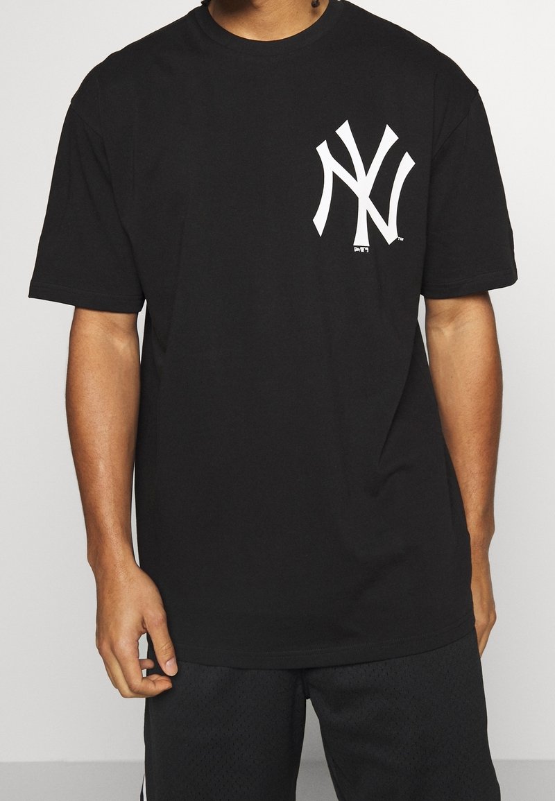 Man med svart oversized t-shirt med en stor vit "NY"-logotyp på vänster bröst och svarta mesh-shorts.