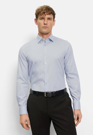 Man die een lichtblauw overhemd met lange mouwen draagt, ingestopt in een zwarte broek met een zwarte riem, staand tegen een effen achtergrond.