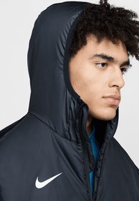 Schwarze Nike-Puffermantel mit hohem Kragen und Reißverschluss, der ein weißes Logo aufweist. Der Stoff wirkt glatt und leicht glänzend.