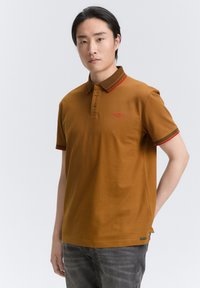 Camisa polo marrón de manga corta, con cuello a rayas en rojo y verde oliva contrastantes, y un pequeño logo rojo en el pecho. Textura suave.