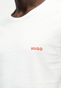 Nahaufnahme einer Person, die ein weißes T-Shirt mit dem Wort „HUGO“ in kleinen orangen Buchstaben auf der linken Brust trägt.