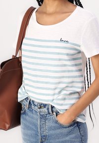 Camiseta blanca de manga corta con rayas azul claro, que presenta un detalle bordado de "amor". Combinada con shorts de denim de cintura alta y una bolsa marrón.