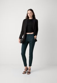 Černý propracovaný blejzr přes černý krátký top, doplněný zelenými slim-fit kalhotami a černými sandály na podpatku. Malá černá crossbody kabelka.