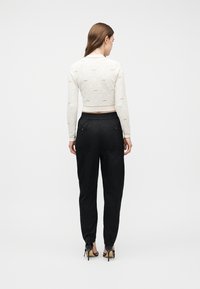 Γυναίκα π ου φοράει λευκό crop top με μακριά μανίκια και σχέδια, μαύρο ψηλόμεσο παντελόνι και μαύρα σανδάλια με ψηλό τακούνι, γυρισμένη με την πλάτη.