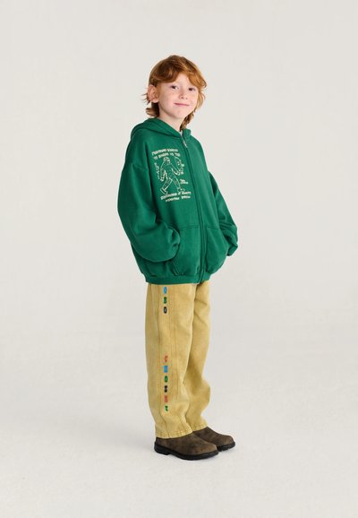 Enfant aux cheveux roux portant un sweat à capuche vert avec un texte graphique et un pantalon beige avec des lettres colorées, debout les mains dans les poches.