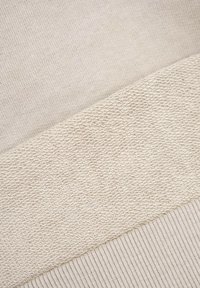 Beżowy materiał z dwiema tkaninami: gładkim dzianinowym i teksturowanym splotem, ze żeberkowym detalem wzdłuż dolnej krawędzi, tworzącym kontrast.