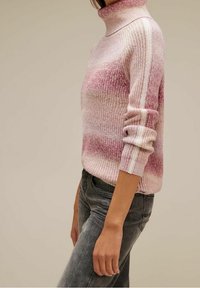 Femme portant un pull en tricot à col roulé rayé rose et blanc à manches longues, assorti à un jean noir délavé, vue de profil.