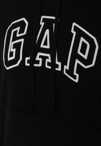 Svart hoodie med vit broderad "GAP" logotyp. Har en känguruficka och dragsko i huvan. Mjukt, texturerat tyg.