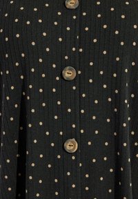 Tissu noir texturé avec des pois crème, comportant cinq grands boutons ronds sur le devant. Conception à côtes verticales sur toute la surface.