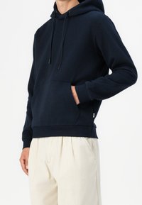 Mörkblå hoodie i mjukt tyg, med en känguruficka, dragsko i huvan och ribbade ärmar. Matchas med ljusbeiga byxor.