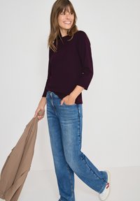 Burgunderfarbener Pullover mit 3/4-Ärmeln, kombiniert mit hellblauen Jeans im geraden Schnitt. Eine beige Jacke in der Hand und weiße Sneakers mit roten Akzenten.