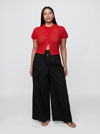 Cardigan rosso lavorato a maglia con maniche corte, orlo a scallop e chiusura a fiocco, abbinato a pantaloni neri a gamba larga e sandali flat.