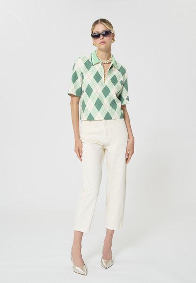 Femme portant un polo à motif losanges vert et blanc, un pantalon blanc cassé, un collier de perles, des lunettes de soleil et des talons argentés, debout devant un fond uni.