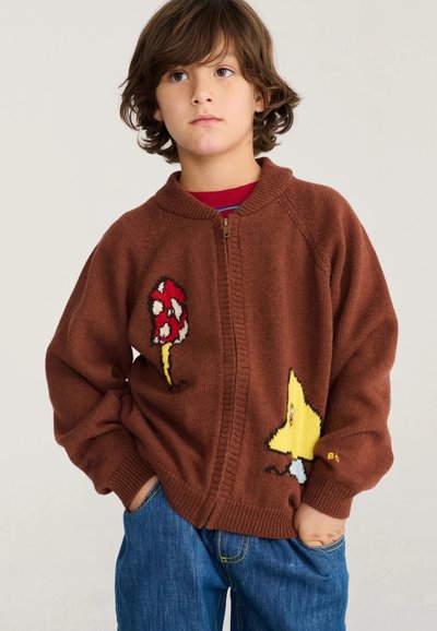 Enfant aux cheveux bruns mi-longs portant un pull à fermeture éclair marron avec des motifs de champignons et d'étoiles et un jean bleu, les mains dans les poches.