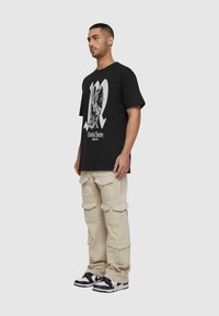 Sort oversized t-shirt med hvid grafisk design og tekst. Beige cargo-bukser med flere lommer og sorte sneakers.