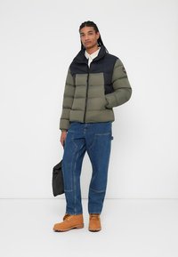 Grøn og sort puffjakke med lynlås, der har et quiltet design. Sammen med blå denimbukser og brune støvler.