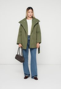 Kvinna klädd i en grön oversized kappa, vit topp, blå vida jeans, håller en mörkbrun handväska, står mot en enfärgad ljus bakgrund.