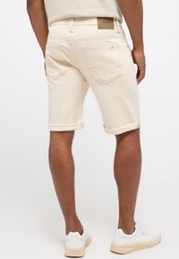 Shorts en denim couleur crème, avec des revers, design à cinq poches et une étiquette en cuir sur la ceinture arrière. Portés avec des baskets blanches.