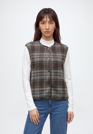 Jeune femme portant un gilet marron à carreaux boutonné sur une chemise blanche à manches longues et un jean bleu, debout devant un fond uni.