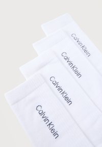 Πέντε ζευγάρια λευκές κάλτσες αστραγάλου με την επωνυμία Calvin Klein, τοποθετημένα σε ένα διαβαθμισμένο αλληλοκαλυπτόμενο μοτίβο πάνω σε ανοιχτό φόντο.