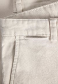 Bež denim tkanina s dijagonalnim rebrima, koja ima petlje za remen i bočni otvor za džep. Tekstura je glatka s blagim sjajem.