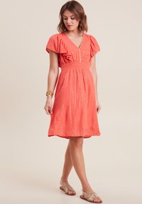 Koralfarbenes kurzes Kleid mit V-Ausschnitt, schwingenden Ärmeln und gerafftem Bund. Verfügt über metallische Fäden und einen fließenden Saum. Trägt dazu goldene Sandalen.