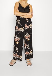 Pantalons larges noirs à motifs floraux avec des fleurs roses et jaunes ; associés à un débardeur noir avec bordure en dentelle. Sandales grises à enfiler avec un accent floral.