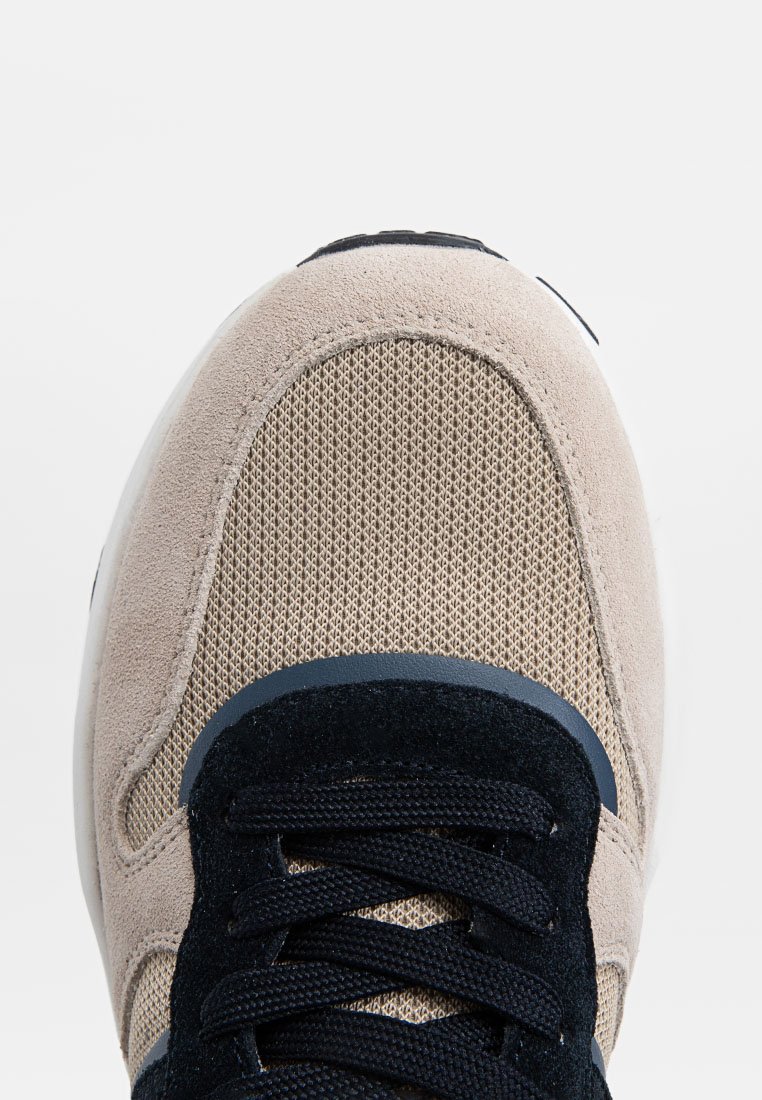 Beige en zwarte sneaker met een mesh neus en suède overlays, met zwarte veters die in een standaard kruisspatroon zijn gestrikt.