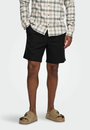Homme portant une chemise à carreaux beige boutonnée, un short noir et des sandales ouvertes couleur beige, debout les mains partiellement dans les poches.