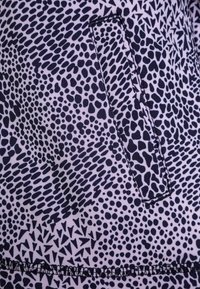 Tkanina w wzory z mieszanką czarnych i fioletowych kształtów, w tym plam i trójkątów. Zawiera małą kieszonkę z widocznym przeszyciem.