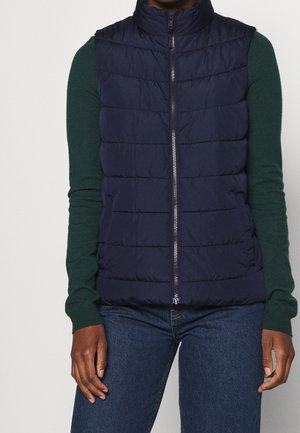 Veste sans manches matelassée bleu marine avec fermeture éclair à l'avant, portée sur un pull vert foncé à manches longues et un jean bleu.