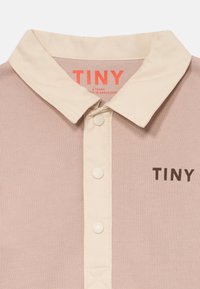 Beige Poloshirt mit cremefarbenem Kragen und Knöpfen, auf der Brust mit "TINY" bedruckt und mit sichtbarem Innenetikett hinter dem Kragen.