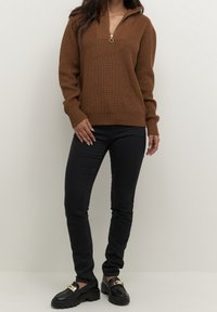 Femme portant un pull en tricot marron avec une fermeture éclair, un pantalon noir ajusté et des mocassins noirs avec des détails dorés, debout devant un fond clair.