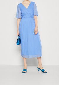 Robe portefeuille bleu clair avec manches courtes et transparentes, jupe fluide et taille cintrée. Associée à des sandales à talons ouvertes bleu sarcelle brillantes et un sac à main.