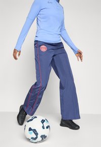 Person som bär en blå långärmad tröja och marinblå Paris Saint-Germain-byxor med röd piping, kontrollerar en vit och blå fotboll.