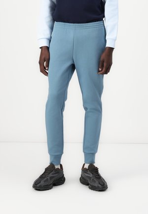 Persona che indossa pantaloni da jogging azzurri abbinati a scarpe da ginnastica nere, in piedi davanti a uno sfondo bianco semplice.