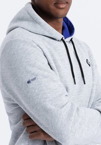 Sudadera de forro polar gris claro con cordones negros y un forro interior azul. Presenta un parche con el logo en el pecho y una etiqueta en la manga.