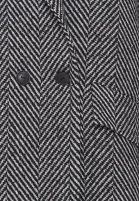 Manteau en laine à chevrons noir et blanc avec deux boutons noirs et une poche à rabat sur le devant.