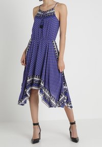 Bezramenné modré midi šaty s potiskem, s lemem ve tvaru kapesníčku, s vázací šňůrkou na krku, s bočními kapsami, nošené s černými sandály na kotníku.