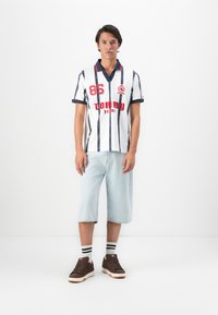 Jonge man in een witte polo met navy strepen en rode letters, lichtblauwe jeanshorts tot de knie, gestreepte sokken en bruine sneakers.