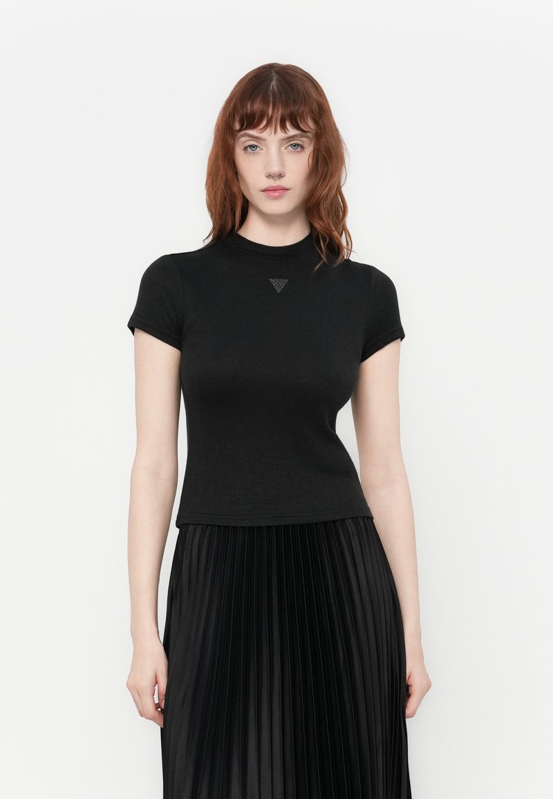 Vrouw met rood haar in een aansluitend zwart shirt met korte mouwen en een zwarte geplooide rok met hoge taille, staande tegen een witte achtergrond.