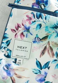 Tejido floral con flores azules, rosas y verdes sobre un fondo blanco. La etiqueta dice "NEXT TAILORING REGULAR FIT" con detalles de la talla.
